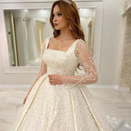 Vestido de novia de lujo con encaje bordado y pedrería, corte princesa, cuello cuadrado, manga larga y botones. 