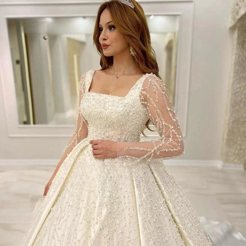 Vestido de novia de lujo con encaje bordado y pedrería, corte princesa, cuello cuadrado, manga larga y botones. 