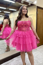 Vestido corto de fiesta de bienvenida con tirantes finos, corte en A, tul escalonado, corsé y apliques en color rosa fucsia.