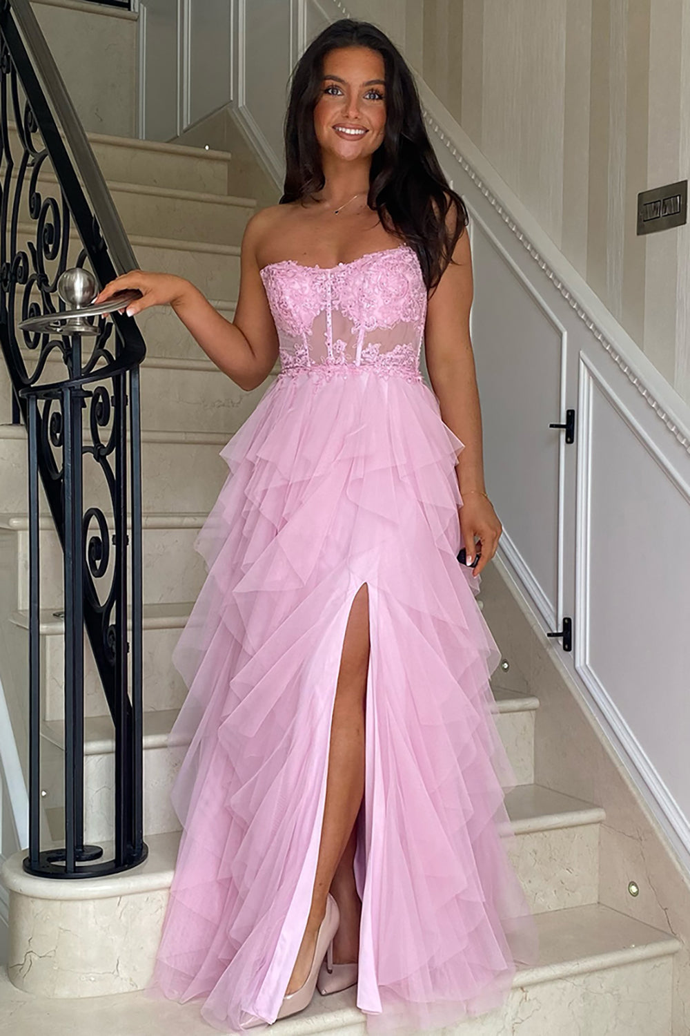 Robe de bal longue bustier en tulle rose poudré, coupe trapèze, à volants et corset orné d'appliques