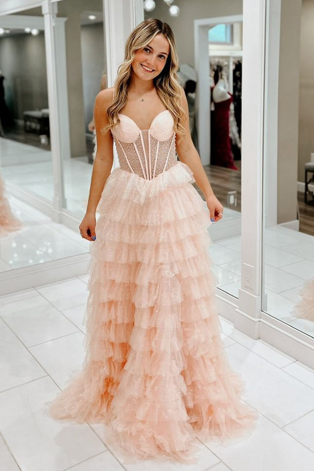 Robe de bal longue trapèze en tulle rose poudré à fines bretelles et à volants, ornée de perles.