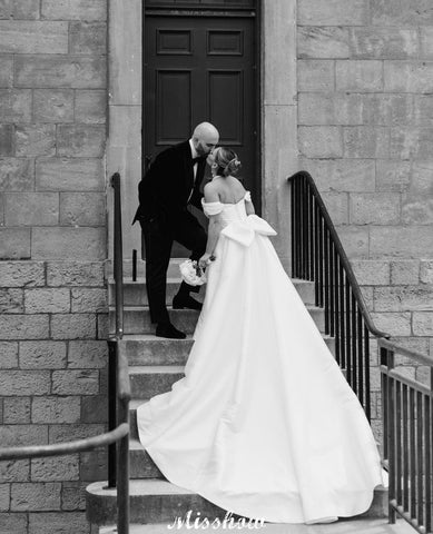 Precioso vestido de novia blanco de corte A, con hombros descubiertos, escote redondo, sin mangas, volantes y abertura frontal en satén.