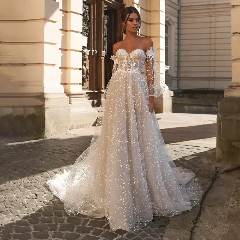 Elegante vestido de novia de corte A, corte sirena, manga larga, hombros descubiertos, escote corazón, tul, apliques de lentejuelas y color beige. 