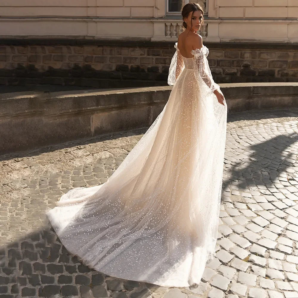 Elegante vestido de novia de corte A, corte sirena, manga larga, hombros descubiertos, escote corazón, tul, apliques de lentejuelas y color beige. 