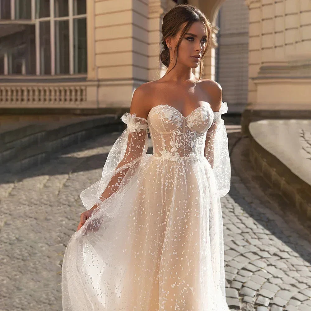 Elegante vestido de novia de corte A, corte sirena, manga larga, hombros descubiertos, escote corazón, tul, apliques de lentejuelas y color beige. 