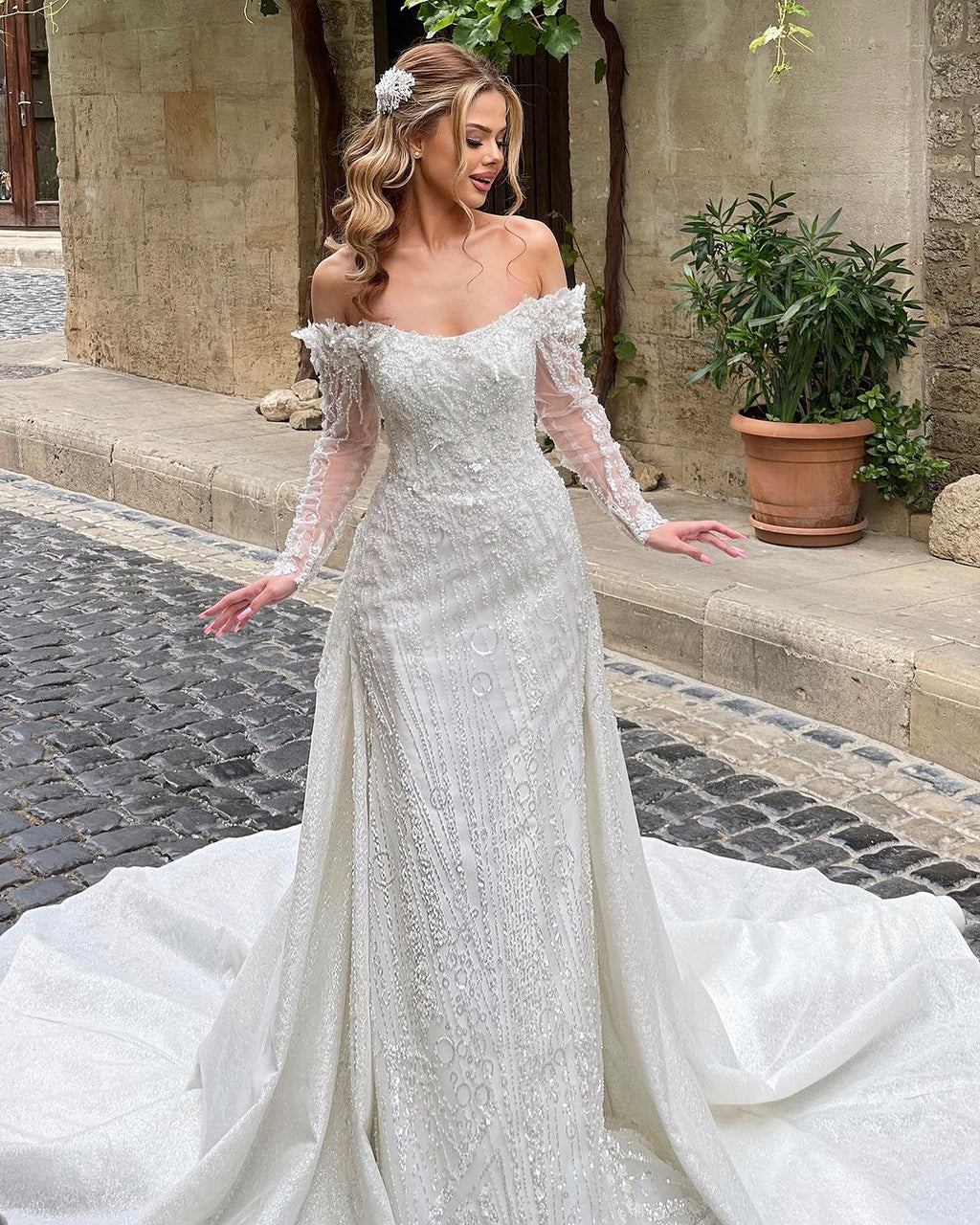 Luxueuse robe de mariée ivoire longue à épaules dénudées, ornée de dentelle et de perles, manches longues et traîne.