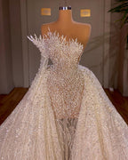 Robe de mariée longue blanche luxueuse, coupe trapèze, ornée de perles et asymétrique à manches.