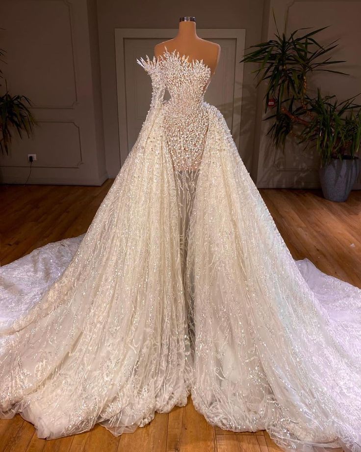 Robe de mariée longue blanche luxueuse, coupe trapèze, ornée de perles et asymétrique à manches.