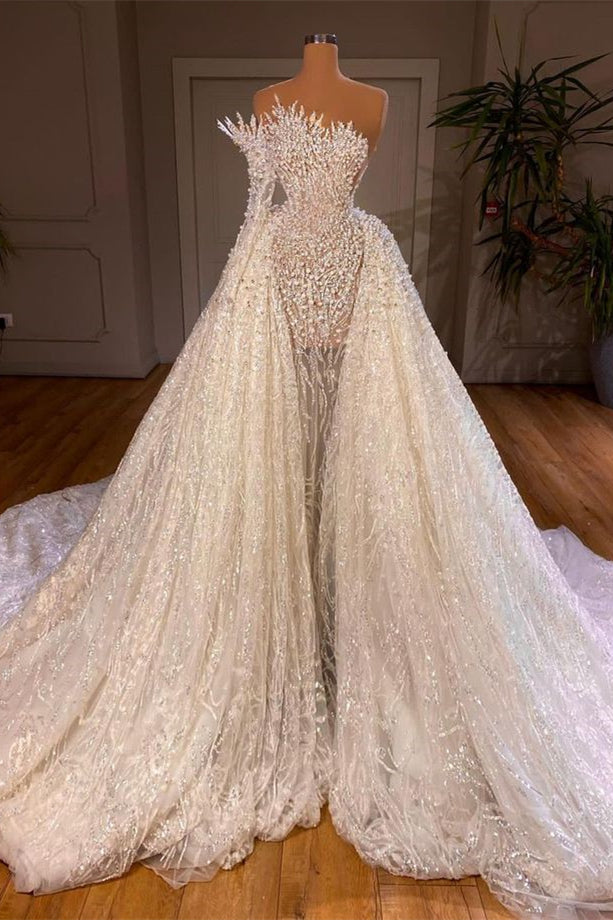 Robe de mariée longue blanche luxueuse, coupe trapèze, ornée de perles et asymétrique à manches.