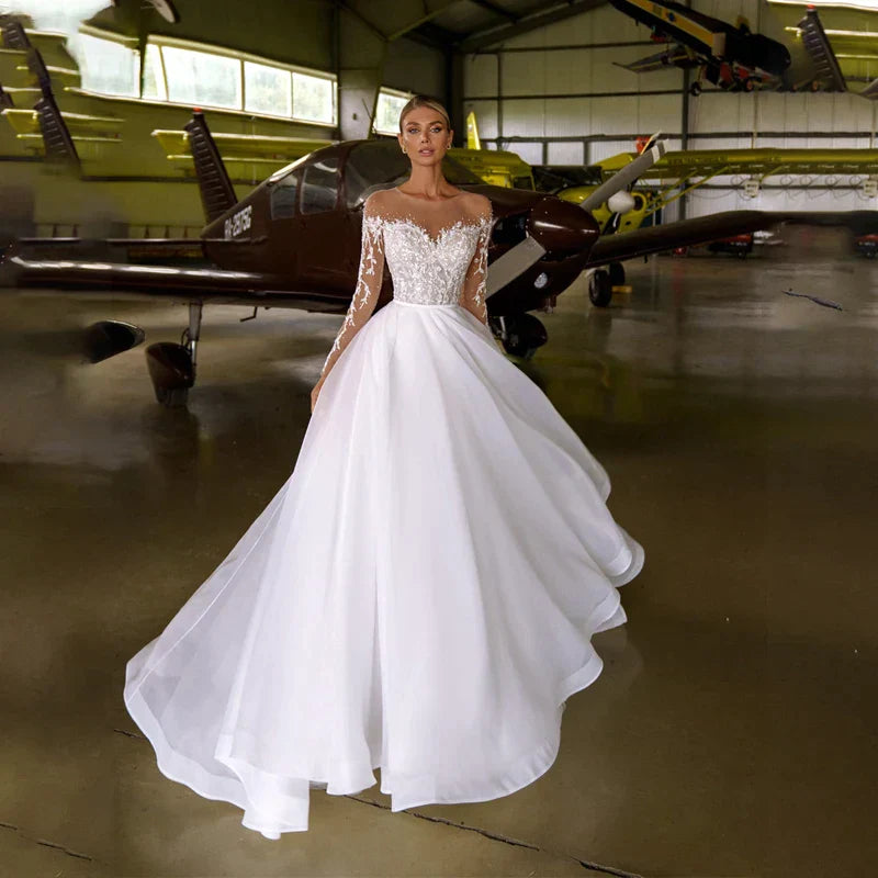 Robe de mariée blanche en organza et tulle, coupe trapèze, ornée de cristaux et à manches longues, avec col transparent. 