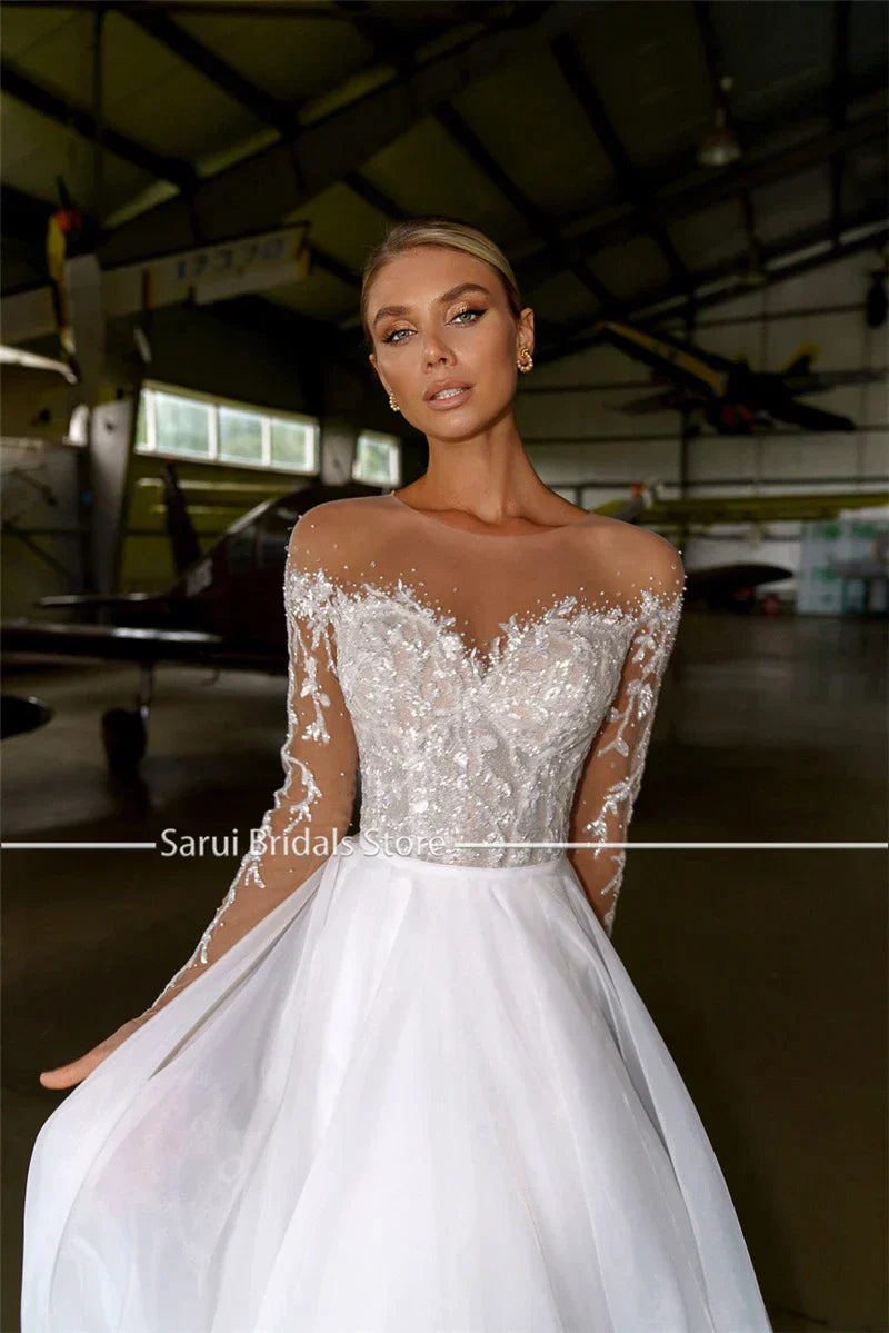 Robe de mariée blanche en organza et tulle, coupe trapèze, ornée de cristaux et à manches longues, avec col transparent. 