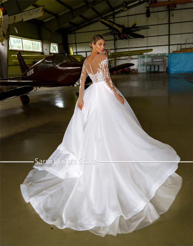 Robe de mariée blanche en organza et tulle, coupe trapèze, ornée de cristaux et à manches longues, avec col transparent. 