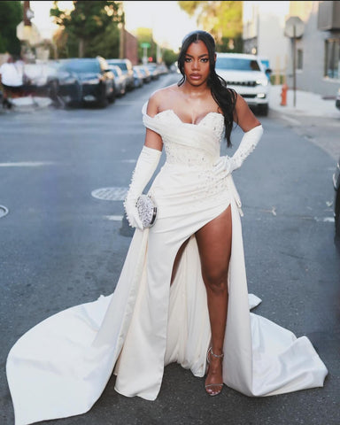 Precioso vestido de novia blanco de corte columna, de satén, sin mangas, con un solo hombro y cola con abertura y detalles de pedrería.