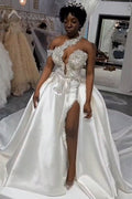 Robe de mariée blanche longue et luxueuse, asymétrique, sans manches, ornée de bijoux et avec traîne amovible.